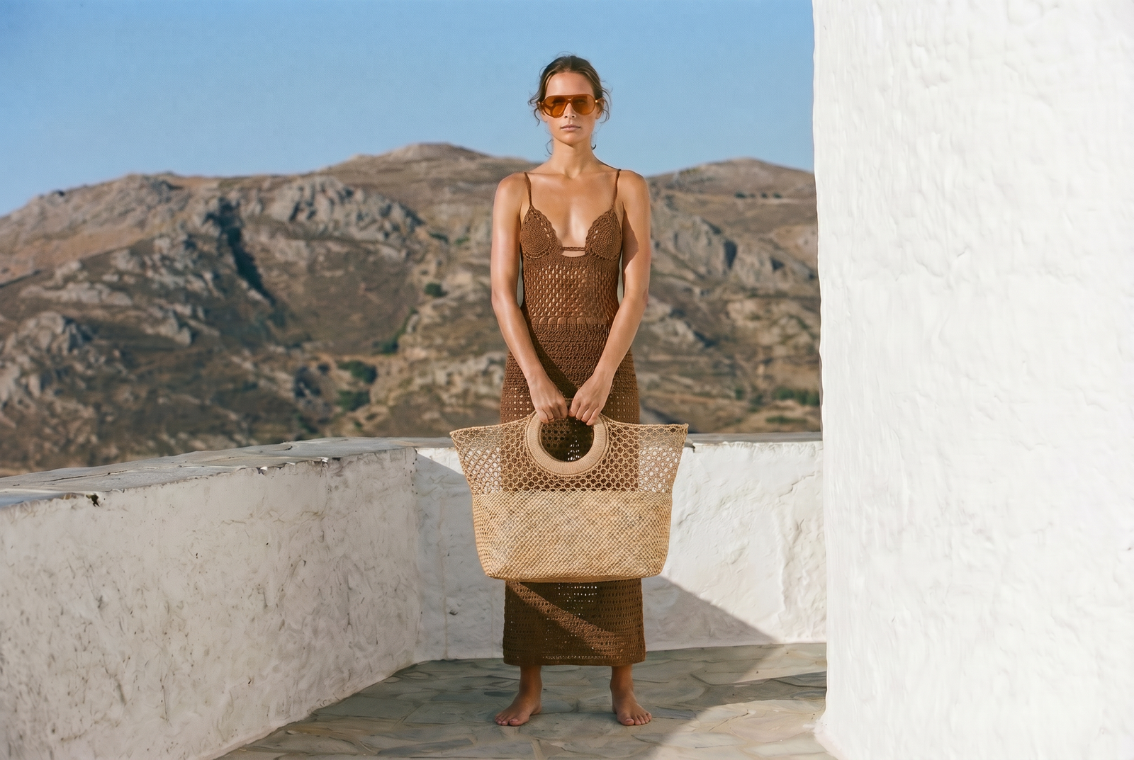 A woman stands barefoot on a white-walled balcony with mountains and blue sky in the background, wearing a brown sleeveless knit dress and holding a large woven straw tote bag.