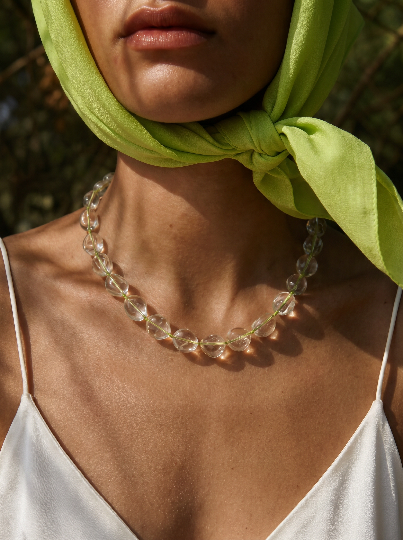 A close-up of a model wearing a chunky clear glass bead necklace with a bright lime green headscarf and a white camisole top.