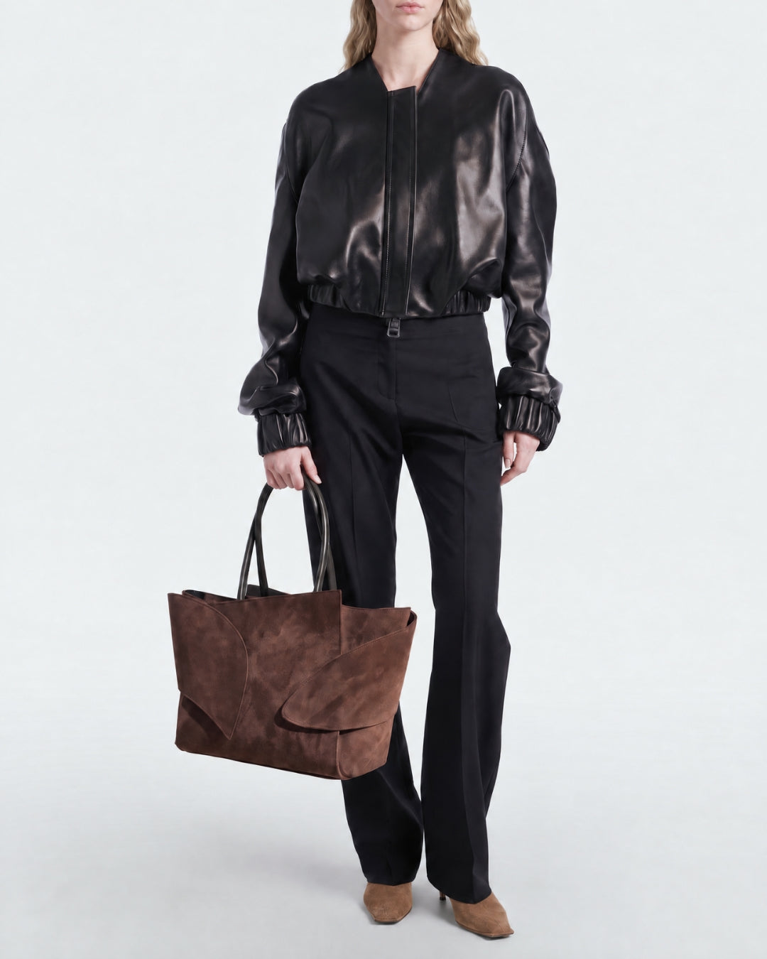 A model wearing a black leather bomber jacket, black wide-leg trousers, and brown pointed-toe heels, holding a brown suede tote bag against a plain light gray studio background.